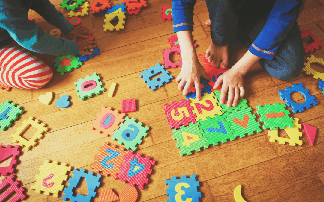 Children playing with colorful foam puzzle pieces and letters scattered on a wooden floor; a child wearing striped pants builds a mat with letters and shapes.