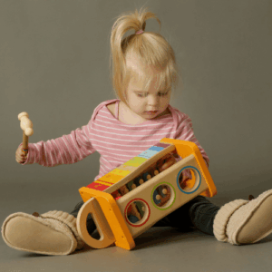Young girl in a pink striped shirt sits on the floor, tapping a wooden mallet against a bright multi-color activity toy.
