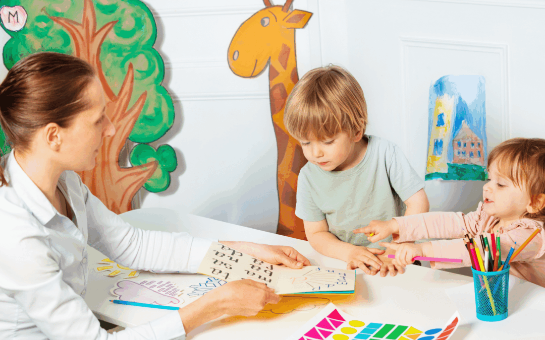 Teacher assists two young children as they cut and paste colorful shapes at a white table in a bright classroom with wall drawings and crafts nearby.